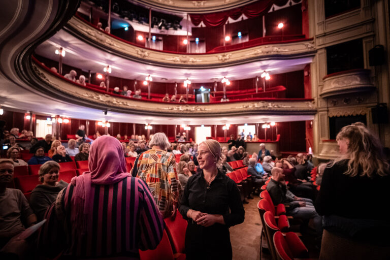Een druk theater met meerdere verdiepingen gevuld met mensen die zich voorbereiden op een voorstelling.