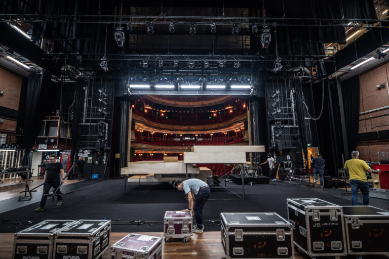 Een team werkt backstage aan de opbouw van een theaterproductie met verlichtingsapparatuur en koffers.