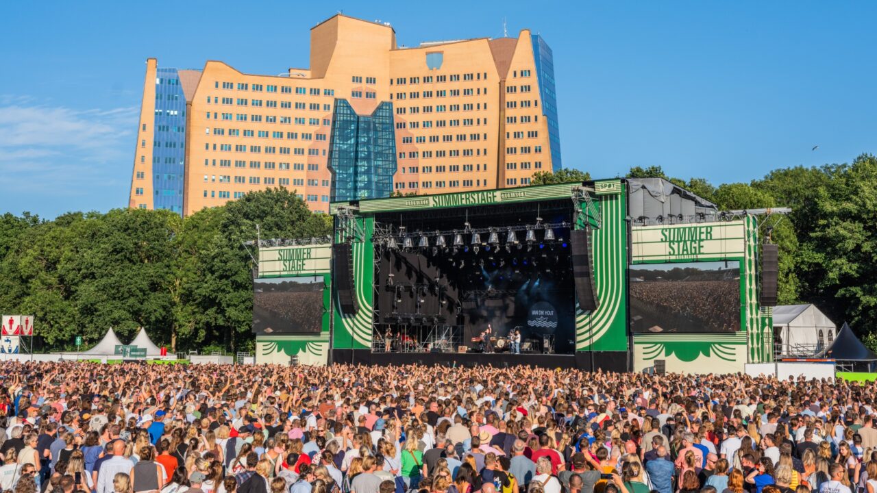 Een druk outdoor muziekfestival met een groot podium en een levendig publiek, tegen een blauwe lucht en een opvallend gebouw op de achtergrond.