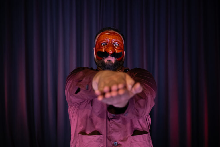 Een man in een paarse outfit met een opvallend rood gezichtsmasker steekt zijn handen naar voren.