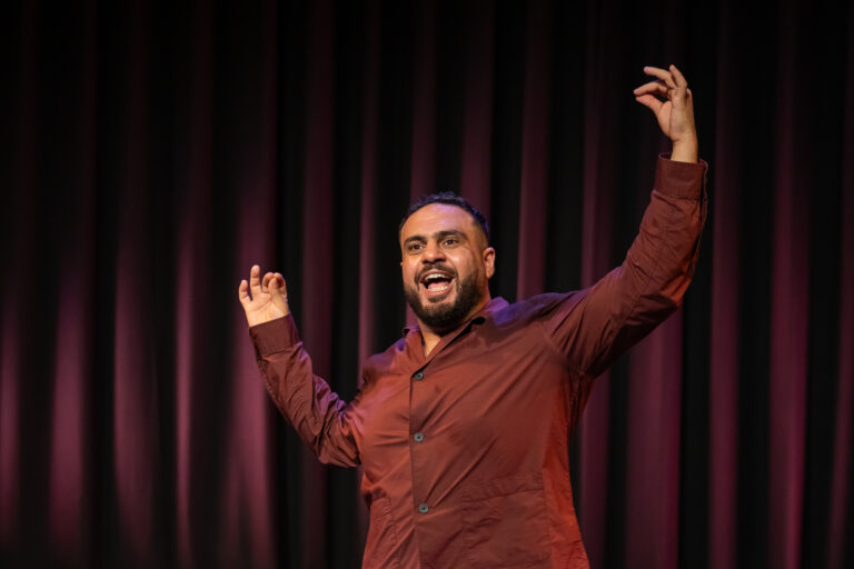 Een performer staat enthousiast op het podium voor een paarse achtergrond en heeft zijn armen omhoog.