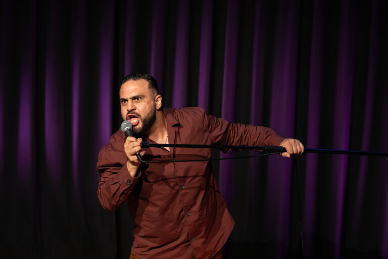 Een man staat op een podium met een microfoon en lijkt enthousiast te praten of te zingen.