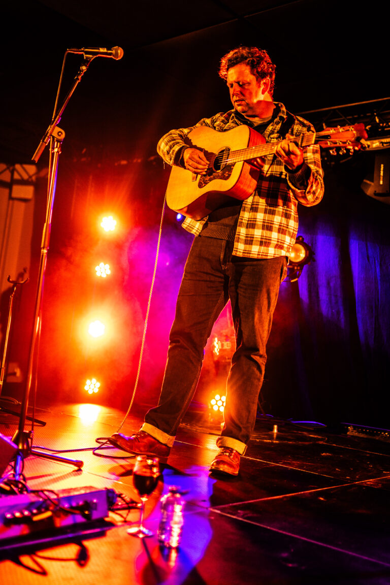Een muzikant staat op een podium met een gitaar, omringd door kleurrijke lichten.