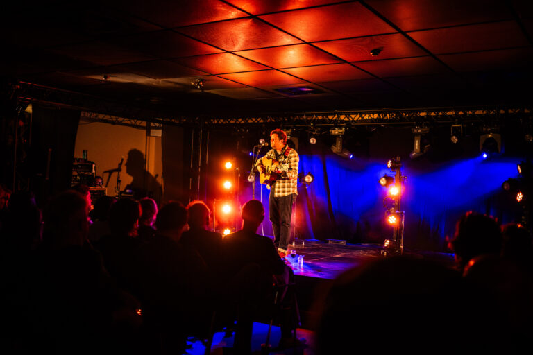 Een comedian staat op een podium in een sfeervol verlichte zaal, terwijl het publiek aandachtig kijkt.