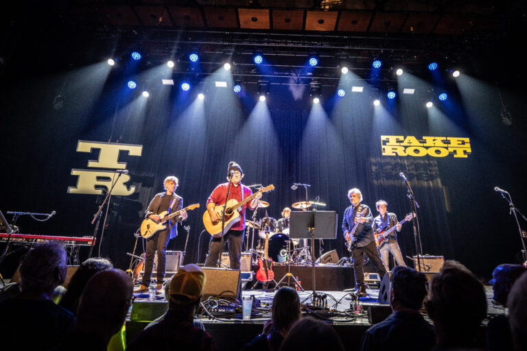 Een band voert live op een podium uit, met muzikanten die verschillende instrumenten bespelen en kleurrijke lichten in de achtergrond.