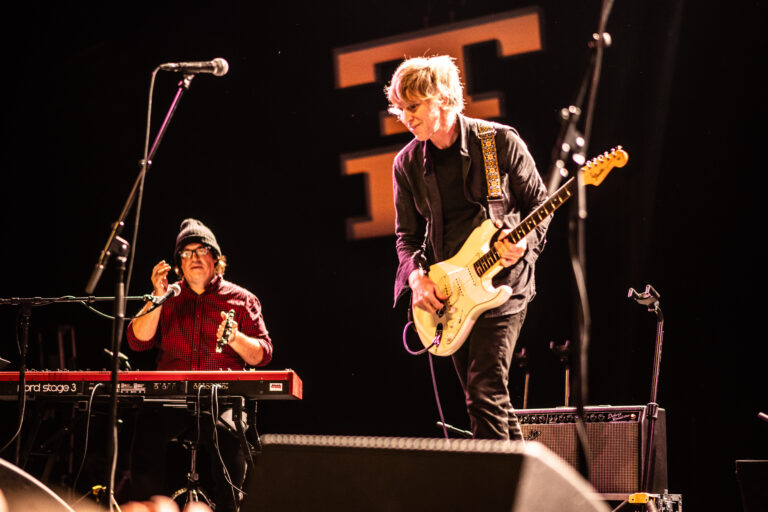 Twee muzikanten staan op een podium, één speelt op een keyboard en de ander op een elektrische gitaar.