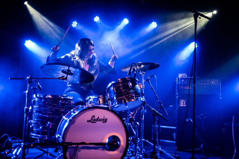 Een drummer speelt op een drumstel terwijl blauwe lichten op het podium stralen.