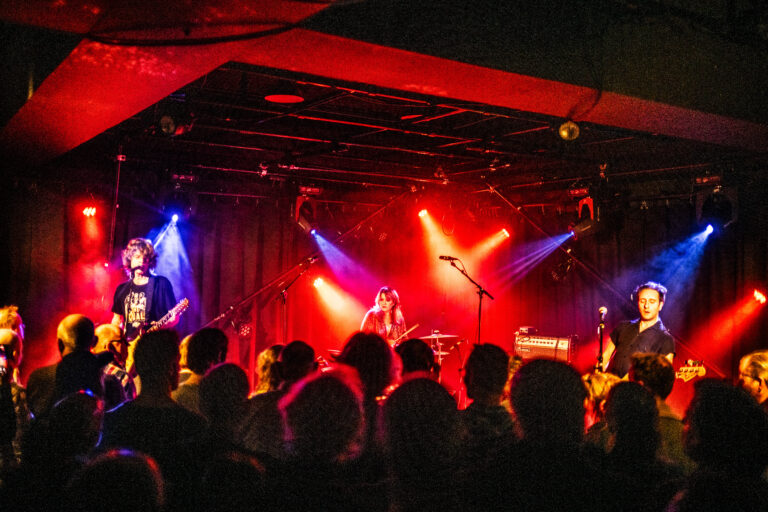 Een band speelt live op het podium, omringd door een publiek onder kleurrijke lichten.