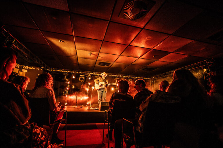 Een muzikant speelt op een podium in een sfeervolle, donkere zaal met een publiek dat aandachtig kijkt.