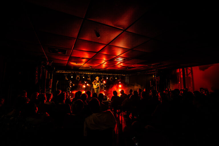 Een live optreden met een muzikant op het podium, omringd door een donker publiek en sfeervolle rode verlichting.