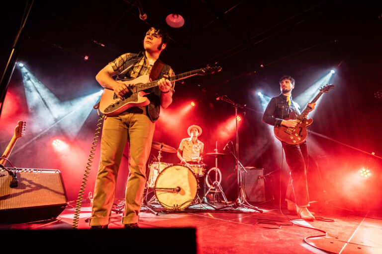 Een band speelt live op het podium met een drummer en twee gitaristen, omringd door kleurrijke ontdekkingslichten.