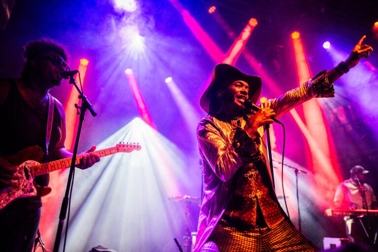 Een energiek optreden van een muzikant in een glanzend kostuum met een gitarrist op de achtergrond, verlicht door kleurrijke lichten.