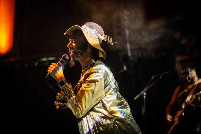 Een artiest zingt enthousiast in een glanzende outfit en hoed, met een microfoon in de hand en een muzikant op de achtergrond.