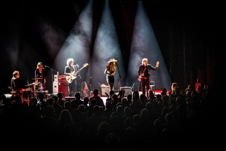 Een band speelt live op een podium voor een enthousiast publiek, omringd door felle lichten.