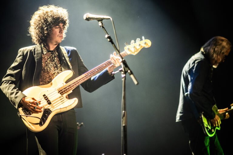 Een muzikant speelt basgitaar op het podium terwijl een andere gitarist op de achtergrond staat.