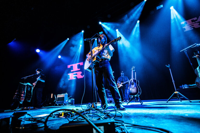 Een muzikant staat op het podium met een gitaar, omgeven door blauwe lichten en andere bandleden in de achtergrond.