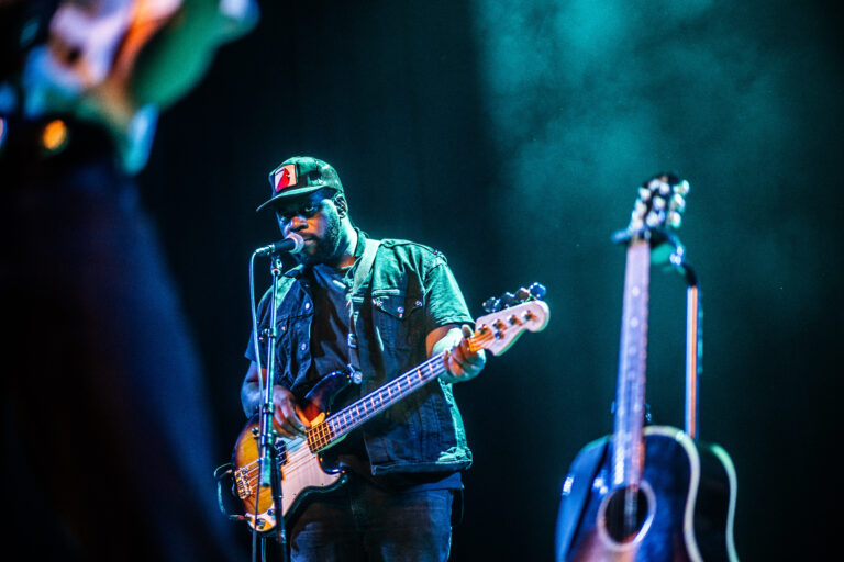 Een muzikant speelt op een elektrisch voorwerp terwijl hij zingt, met een akoestische gitaar op het podium naast hem.