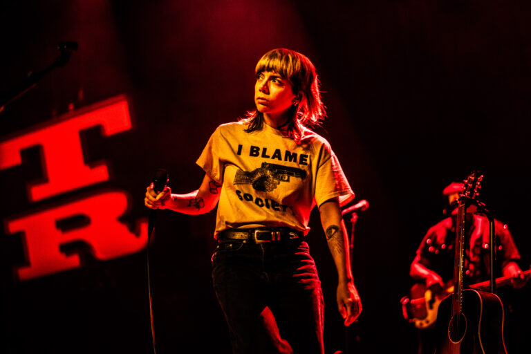 Een muzikante staat op het podium in een opvallend licht, terwijl ze een T-shirt met de tekst 'I BLAME IGOR' draagt.