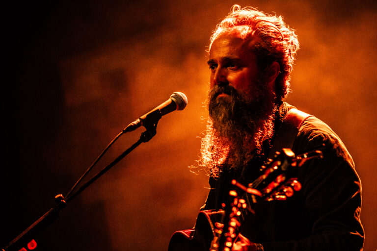 Een muzikant met een volle baard, die een gitaar vasthoudt en zingt voor een microfoon, met een warme oranje achtergrond.