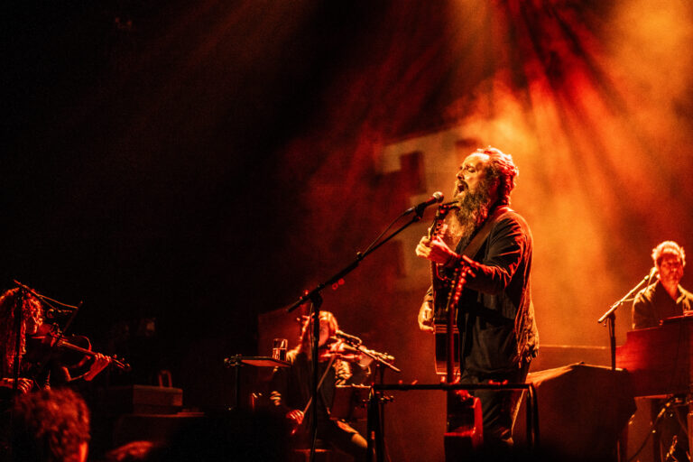 Een muzikant zingt en speelt gitaar op een podium, omringd door andere bandleden, met een dramatische lichtshow op de achtergrond.