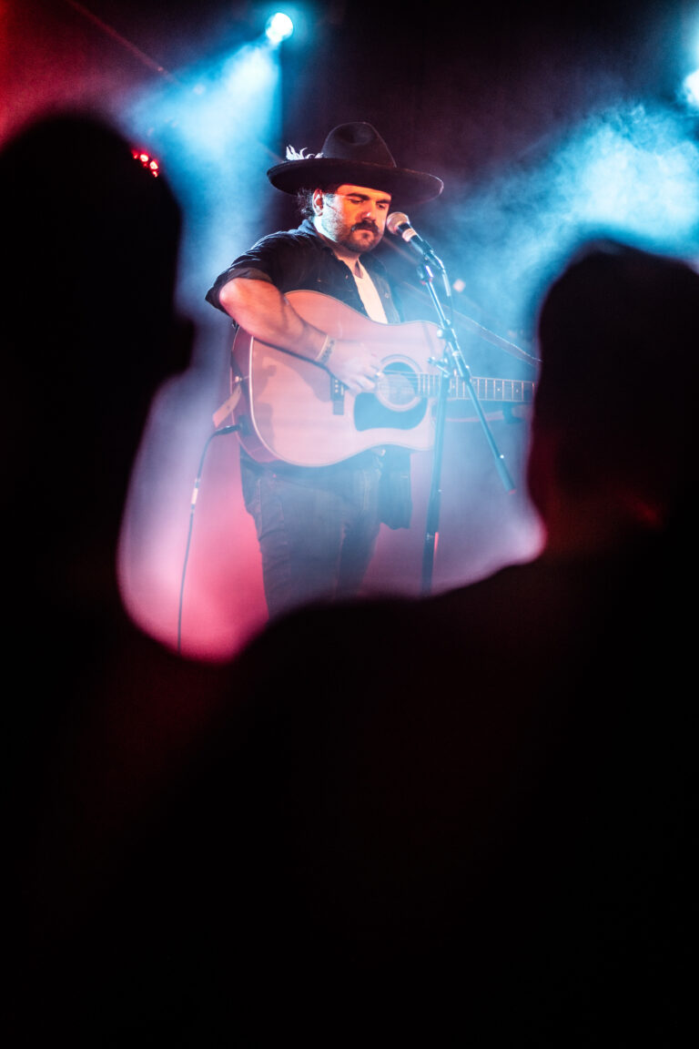 Een muzikant met een gitaar zingt op het podium, omgeven door kleurrijke lichteffecten en rook.