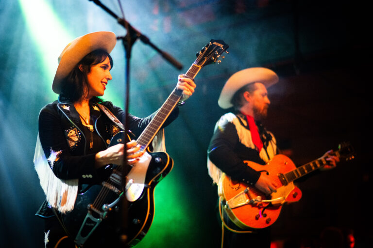Twee muzikanten in westernstyle kleding spelen gitaar op een podium, omringd door kleurrijke verlichting.