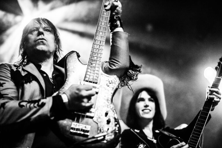 Twee muzikanten, een man met een gitaar en een vrouw die een gitaar vasthoudt, optreden op een podium in zwart-wit.