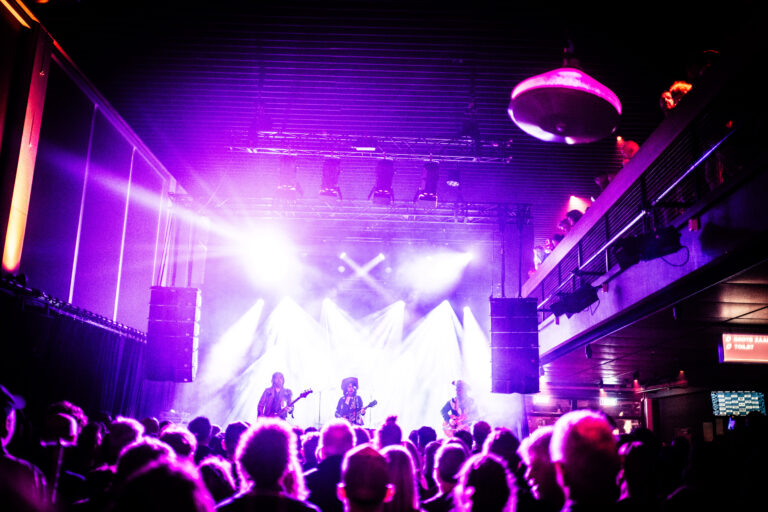 Een liveband speelt op het podium met felle lichten, terwijl het publiek eronder toekijkt.
