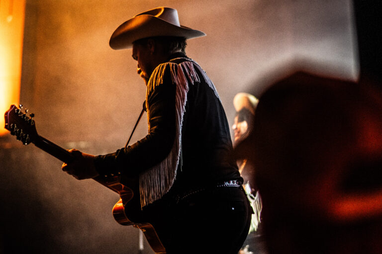 Een muzikant in een cowboyhoed speelt gitaar op een podium, omringd door gespierde lichten en rook.