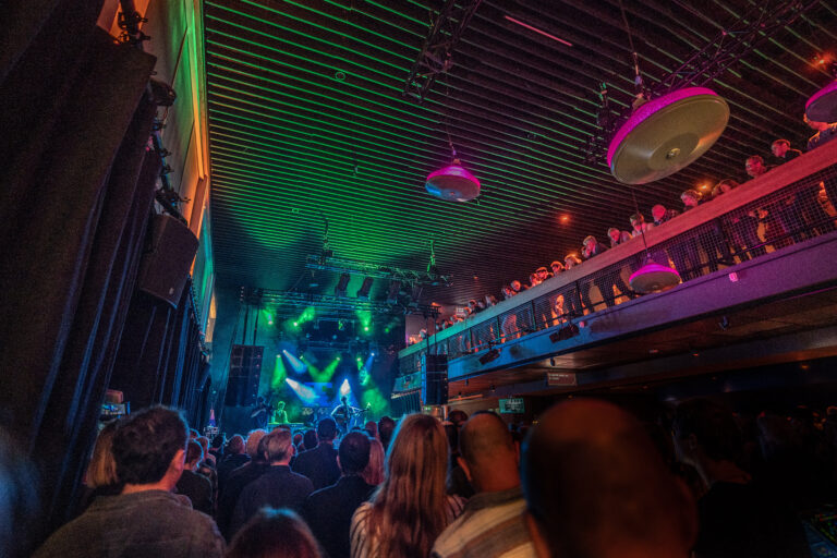 Een drukke muziekshow met een publiek dat naar een podium kijkt, verlicht door kleurrijke lichten.