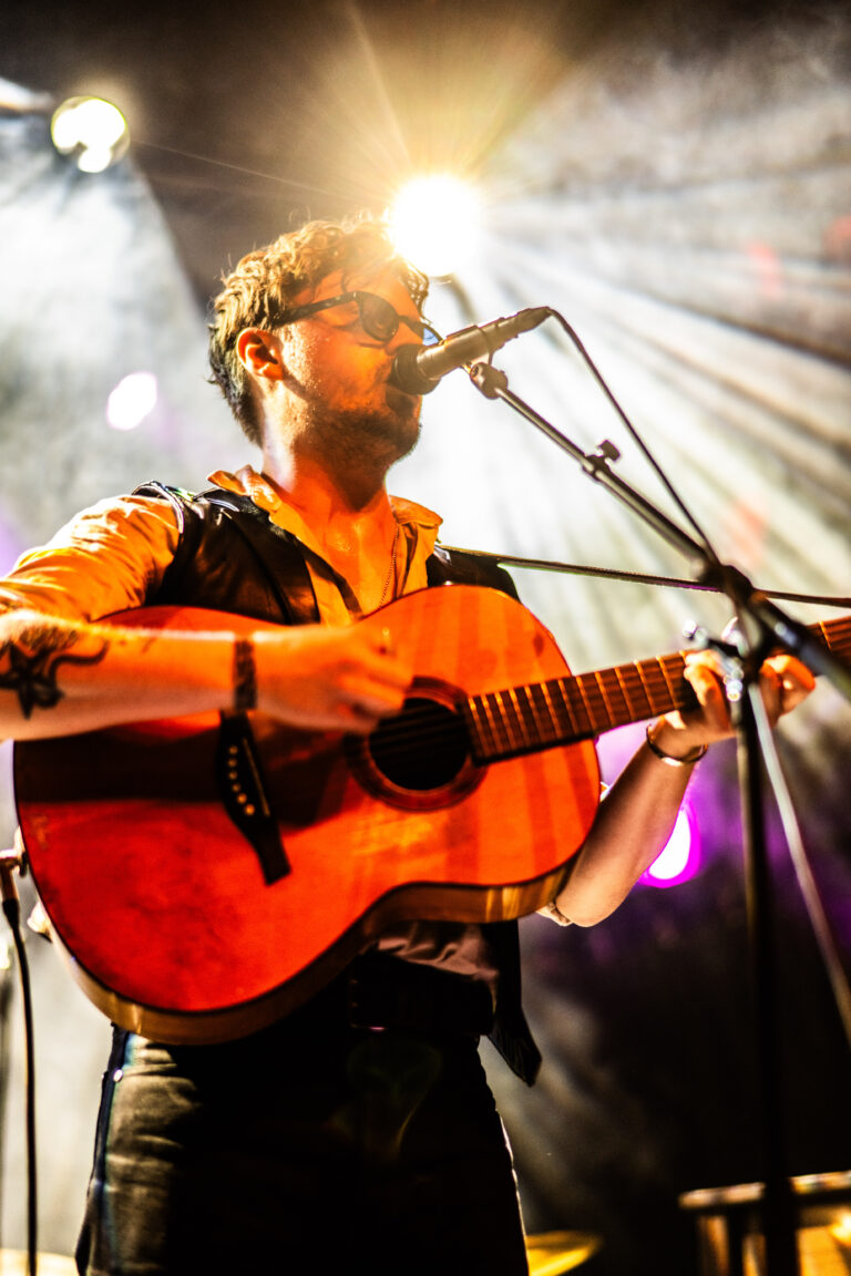 Een muzikant speelt gitaar op het podium met kleurrijke lichtstralen op de achtergrond.
