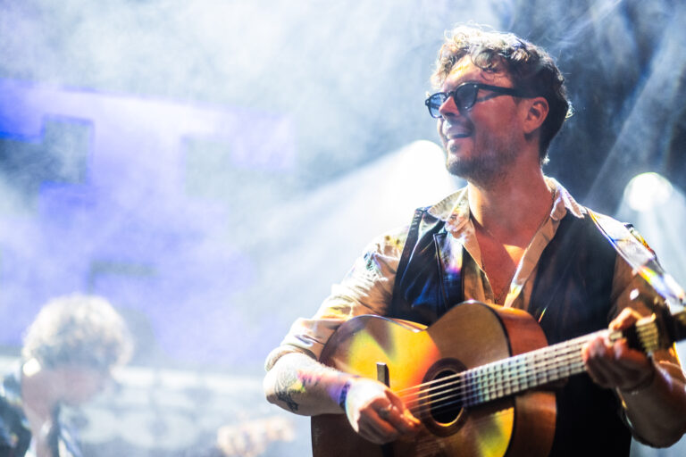 Een muzikant speelt gitaar op een podium met rook en lichteffecten op de achtergrond.