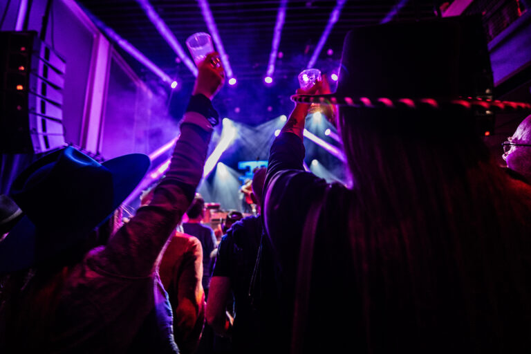 Een groep mensen steekt hun glazen omhoog tijdens een live optreden onder kleurrijke lichten.