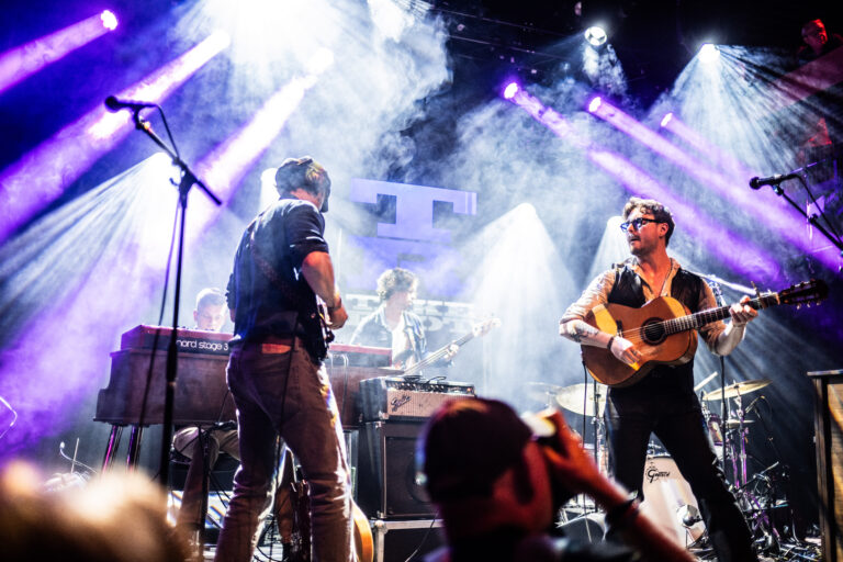 Een band treedt op op een podium met kleurrijke lichteffecten en mist, waarbij muzikanten actief bezig zijn met hun instrumenten.