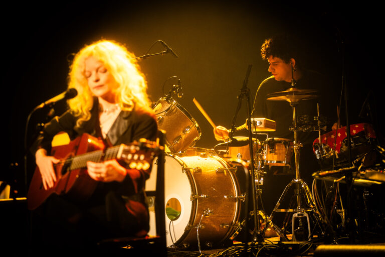 Een muzikant met een gitaar zingt terwijl een drummer op de achtergrond speelt, omringd door sfeervolle belichting.