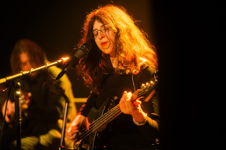 Een muzikante speelt gitaar en zingt tijdens een optreden, met warme belichting op het podium.