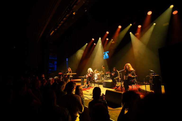 Beeld van een live concert met een band die optreedt op een podium, omringd door een enthousiast publiek.