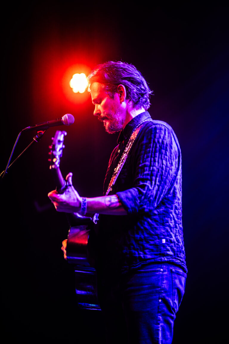 Mann met gitaar op een podium, verlicht door kleurige lichten.