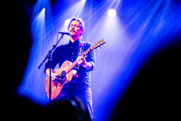 Een muzikant staat op een podium met een gitaar, omringd door blauwe lichtstralen.