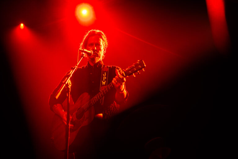 Een muzikant die een gitaar speelt en zingt, verlicht door rode lichten op het podium.