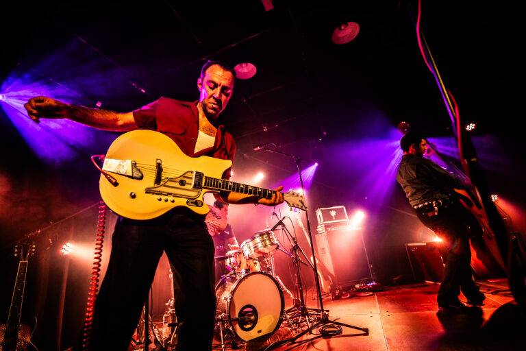 Een muzikant speelt gitaar op een podium met kleurrijke verlichting en een andere bandlid op de achtergrond.