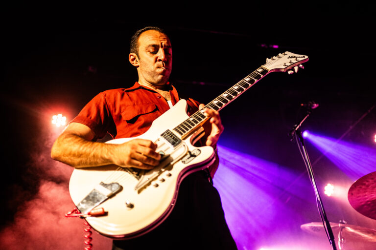 Een muzikant speelt intensief op een witte elektrische gitaar tijdens een optreden met kleurrijke lichten op de achtergrond.