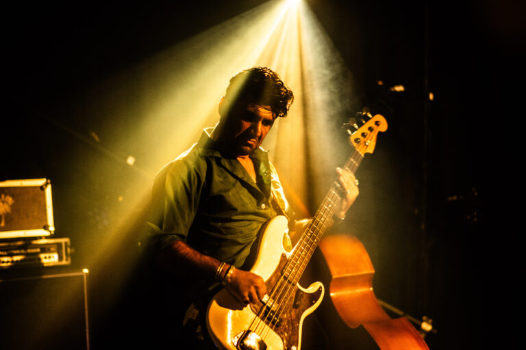 Een muzikant speelt een elektrische basgitaar op een podium, omgeven door sfeervolle lichtstralen.