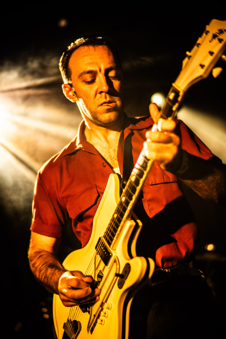 Een muzikant speelt gitaar op een podium, omgeven door dramatische verlichting.