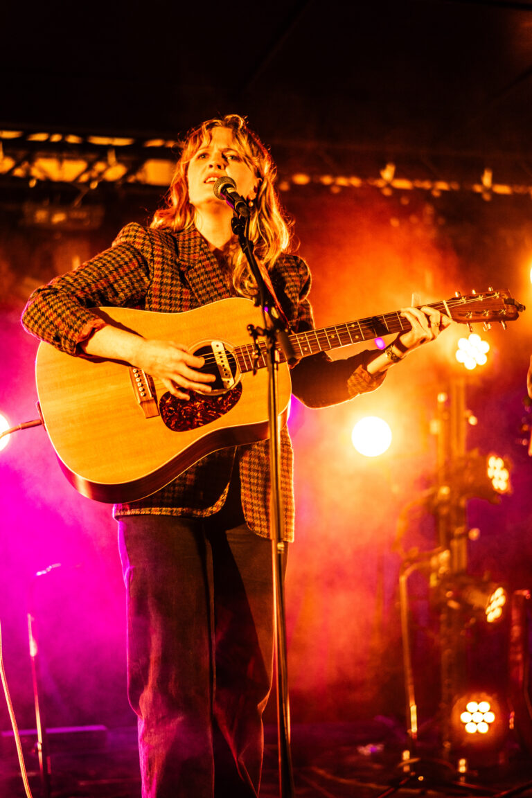 Een muzikant met een gitaar staat op het podium, omringd door kleurrijke verlichting.