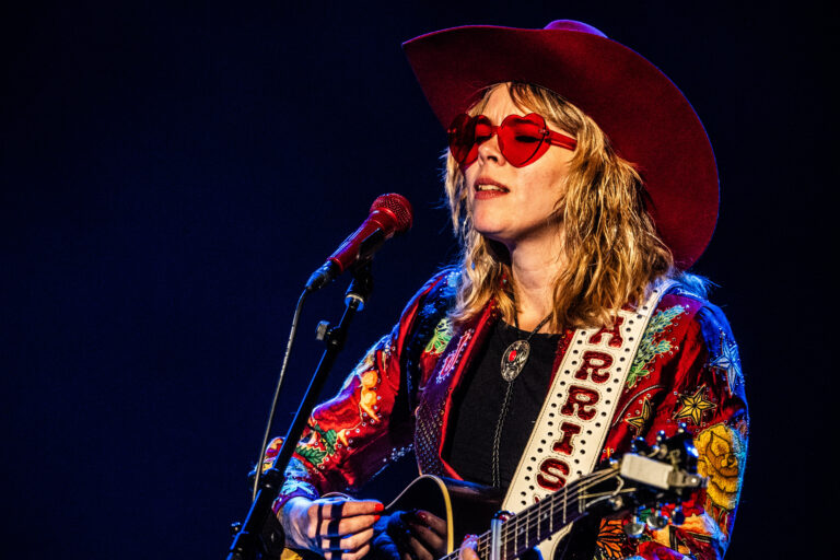 Een muzikante in een kleurrijke outfit en grote hoed zingt terwijl ze gitaar speelt.