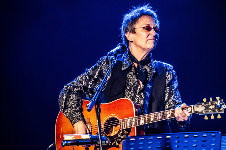 Een muzikant in een zwart-witte outfit, met een gitaar, die op het podium speelt.