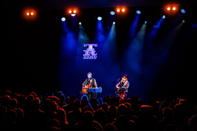 Twee muzikanten treden op voor een publiek, verlicht door kleurrijke lichten op het podium.