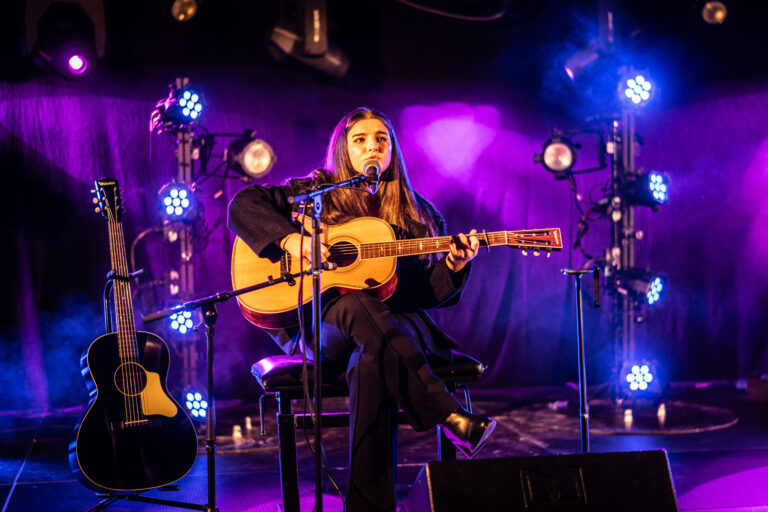 Een muzikante speelt gitaar op podium met paarse verlichting.