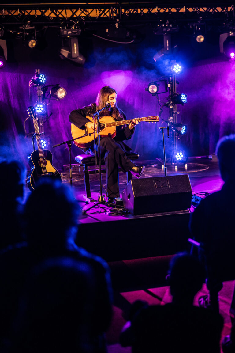 Een muzikant speelt gitaar op een podium met kleurrijke verlichting en publiek op de voorgrond.